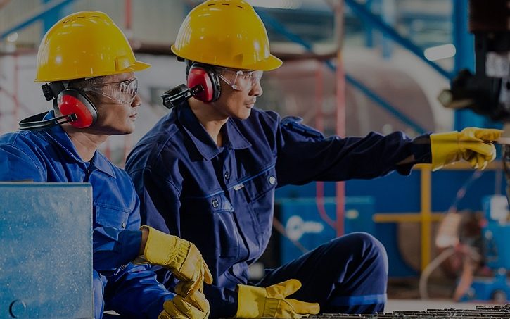 Dos técnicos con casco amarillo y protección auditiva revisan un equipo en planta