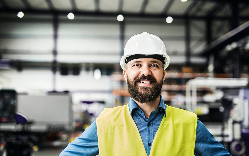 Ingeniero de planta con casco blanco y chaleco reflejante sonriendo en la nave